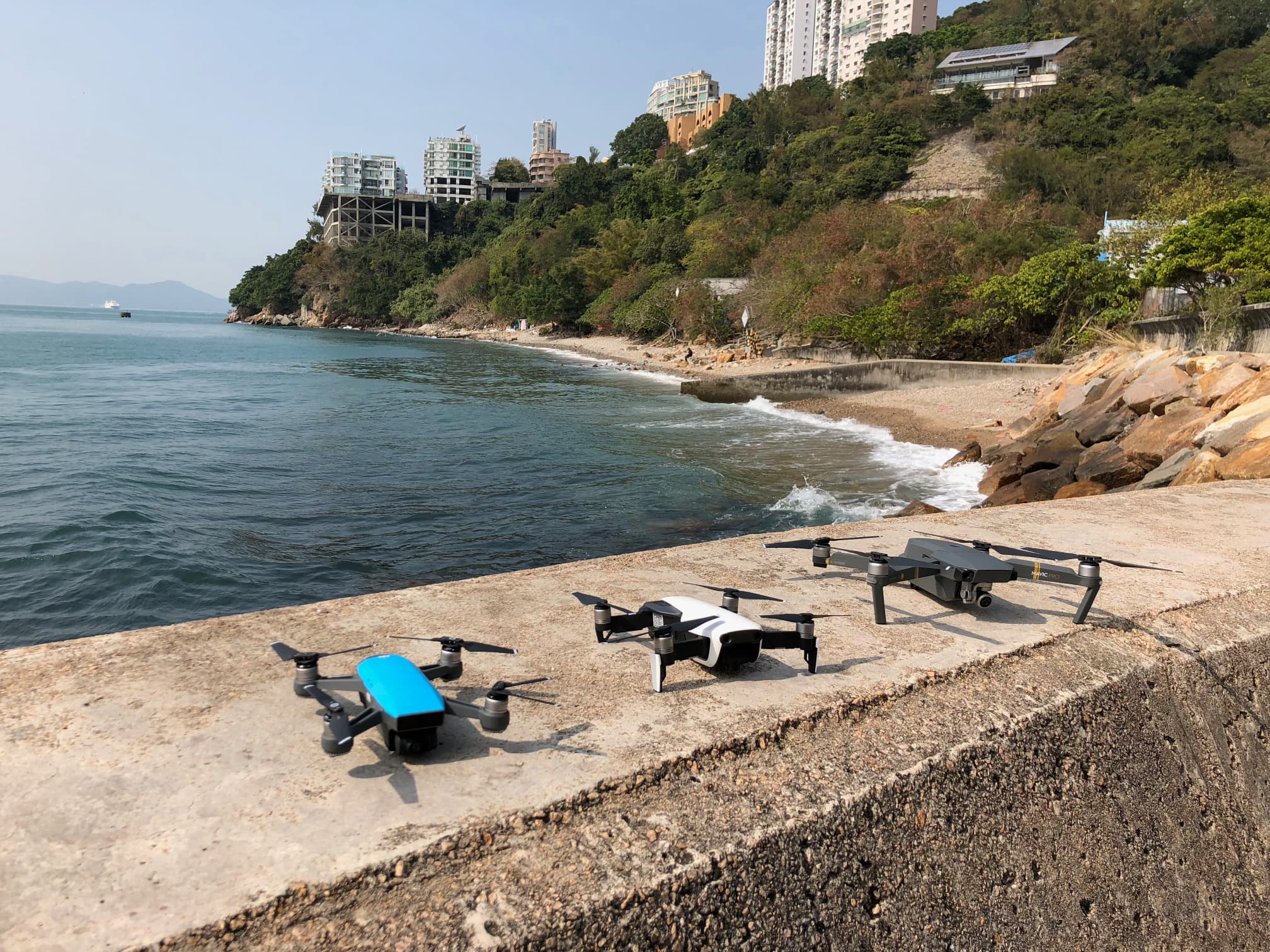 Three DJI drones sit on a sea wall next to a shoreline in Hong Kong.