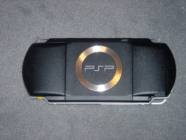 A photo of a PlayStation Portable lying face-down on grey carpet. The PSP logo is visible inside a metallic silver circle.